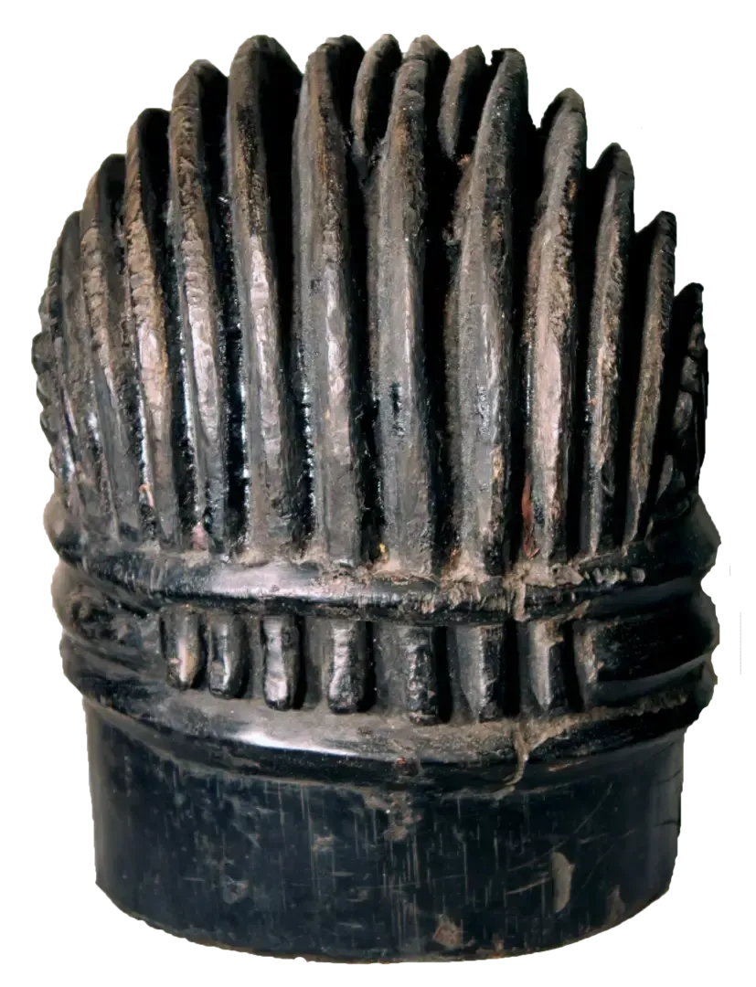 A dark, carved wooden head or mask piece featuring vertical ridges topped by a decorative horizontal band.
