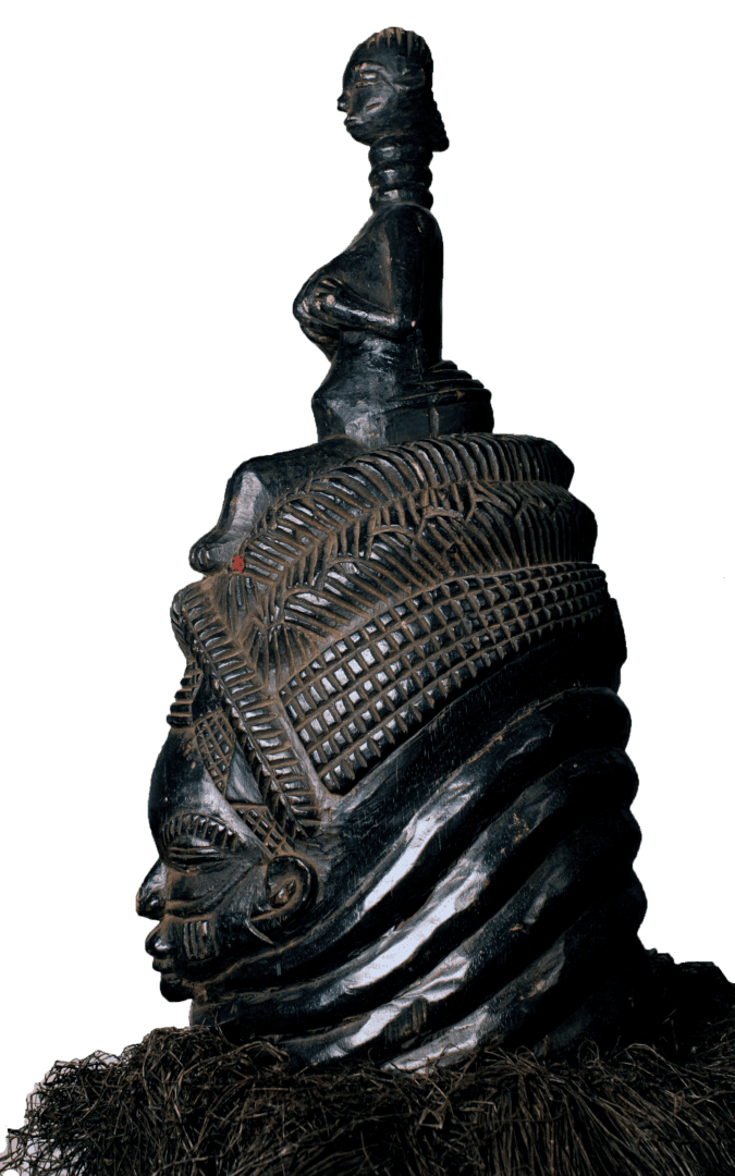 A dark, intricately carved wooden mask featuring a smaller figure on top, ornate hair, and a raffia base.
