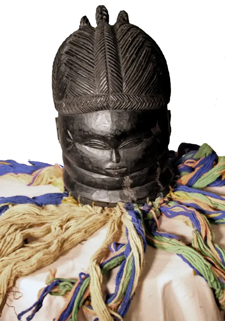 A carved wooden mask with stylized hair, ringed neck, and closed eyes, surrounded by colorful fabric tassels.