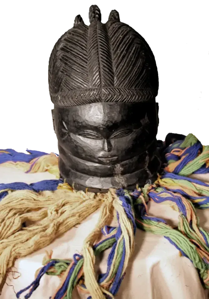 A dark wooden mask with an intricate hairstyle, ringed neck, and colorful fringe, resting on a light-colored surface.