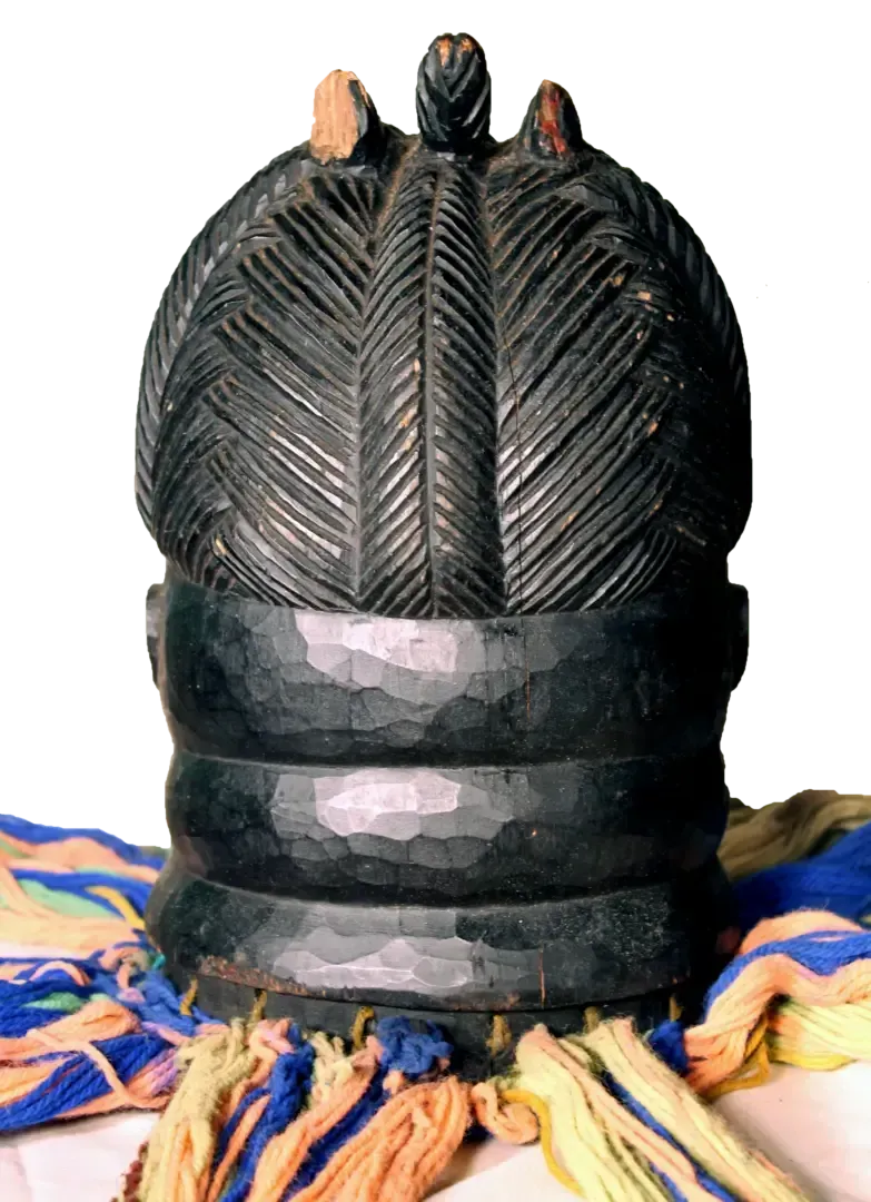 A dark carved wooden helmet mask with deeply textured, etched hair and prominent neck rolls, resting on colorful fabric.