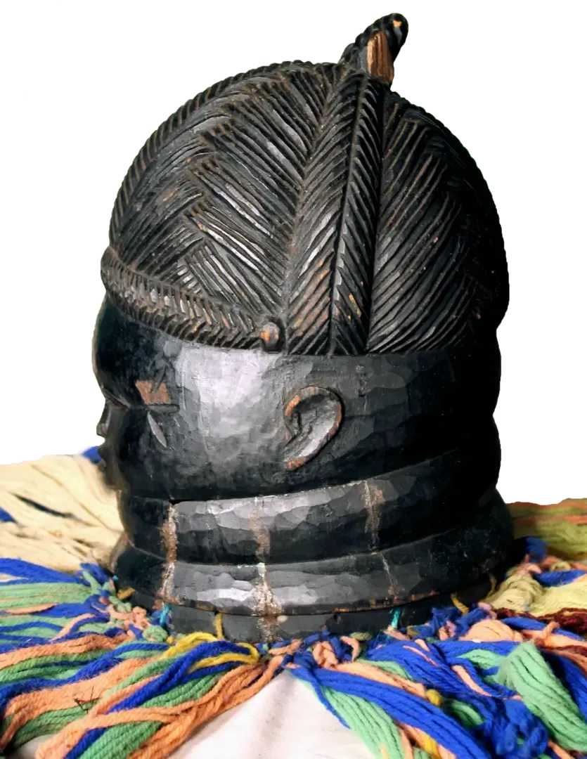 A black, carved wooden helmet mask featuring elaborate hair textures and neck rings, adorned with colorful fabric fringe.
