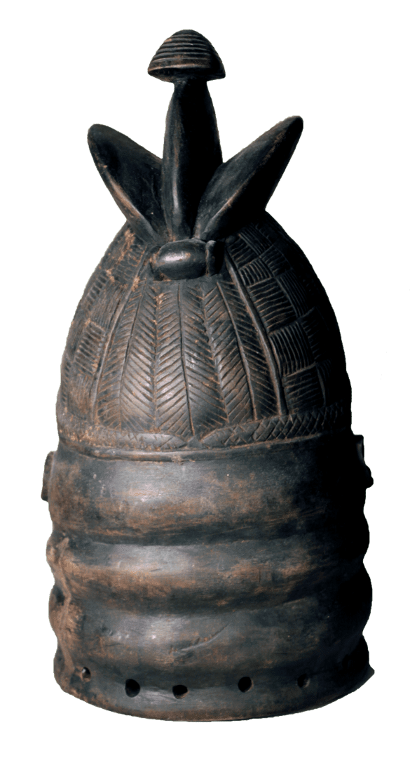 A dark, cylindrical wooden mask with horizontal ridges, patterned carvings on top, and a central finial, on a white ground.
