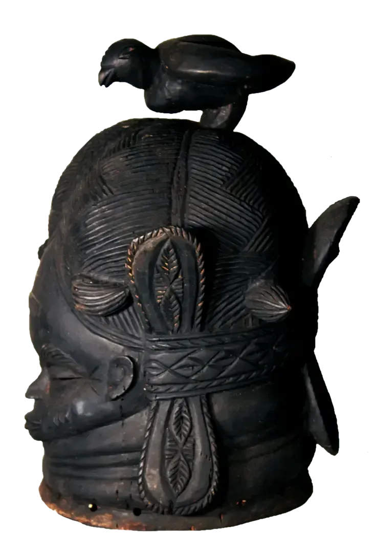 A dark, carved wooden helmet mask featuring an intricate hairstyle with a sculpted bird perched on top.