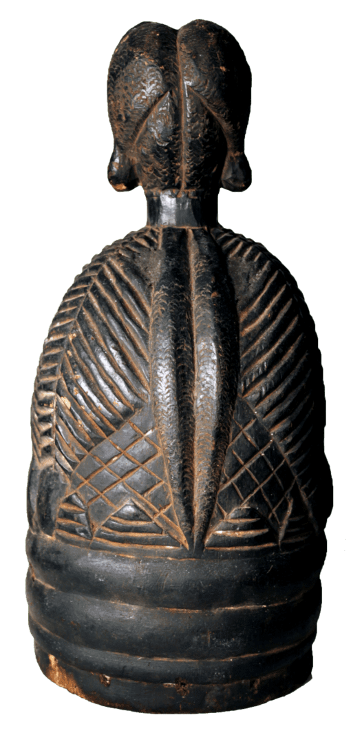 A dark, carved wooden figure with a stylized head, textured geometric patterns on the body, and horizontal base rings.