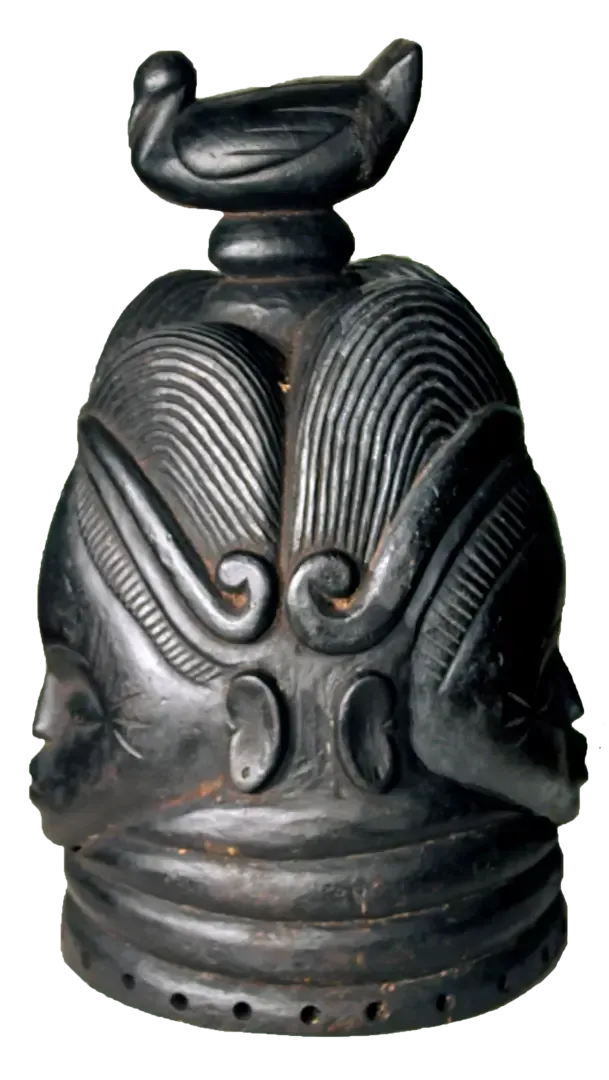 A dark, carved wooden mask with four faces around a central column, topped with a bird figure and ornate, ridged hairstyles.