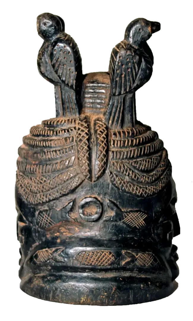 A dark, carved wooden helmet mask featuring two birds perched on top, adorned with detailed patterned textures.