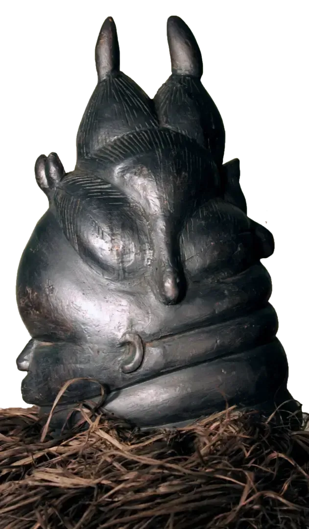A black, carved wooden mask with horn-like protrusions and layered, rounded forms, resting on a base of dried fibers.