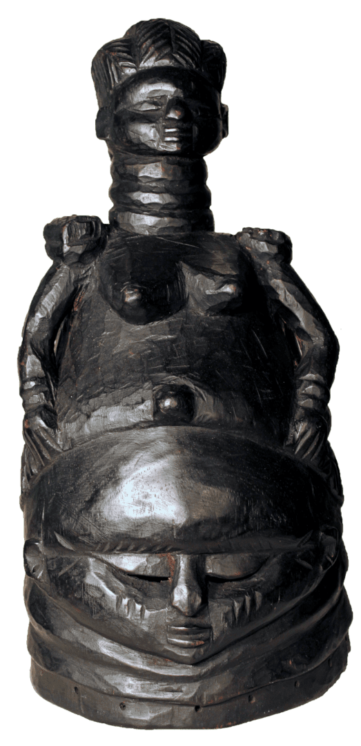 A black, carved wooden sculpture featuring a small female figure seated atop a large, detailed mask with neck rings.