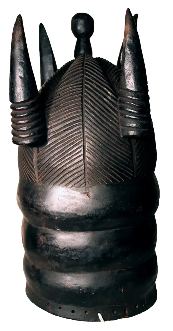 A dark, cylindrical wooden mask with horizontal ridges, carved leaf patterns, and four upward-pointing horn-like spikes.