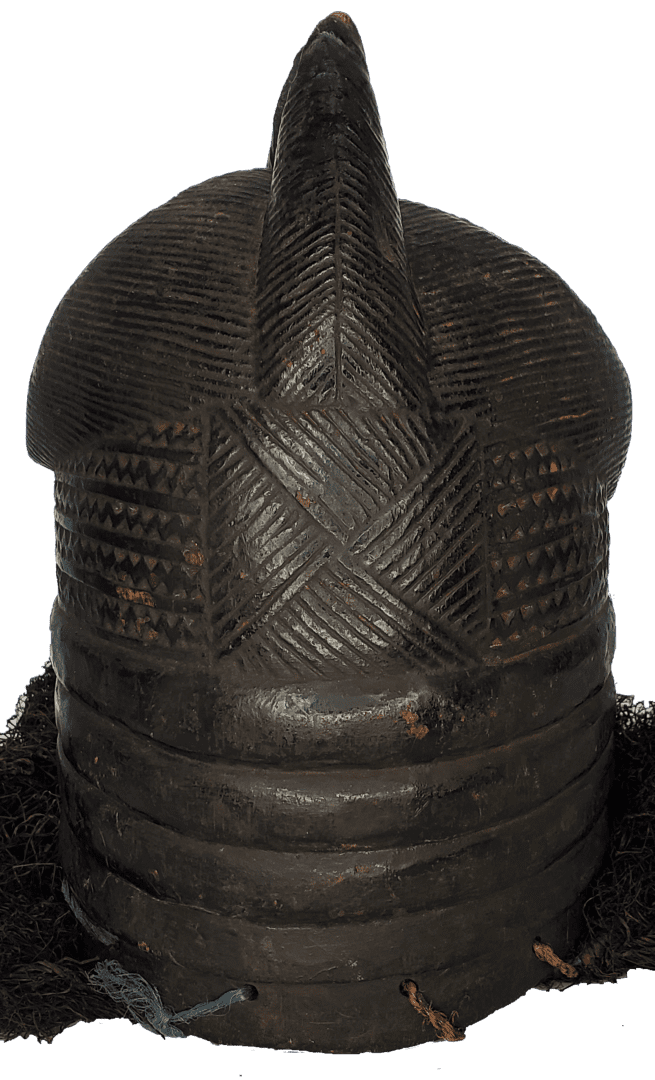 A dark, carved wooden helmet mask with geometric incised patterns, a central crest, and attached dark fiber elements.
