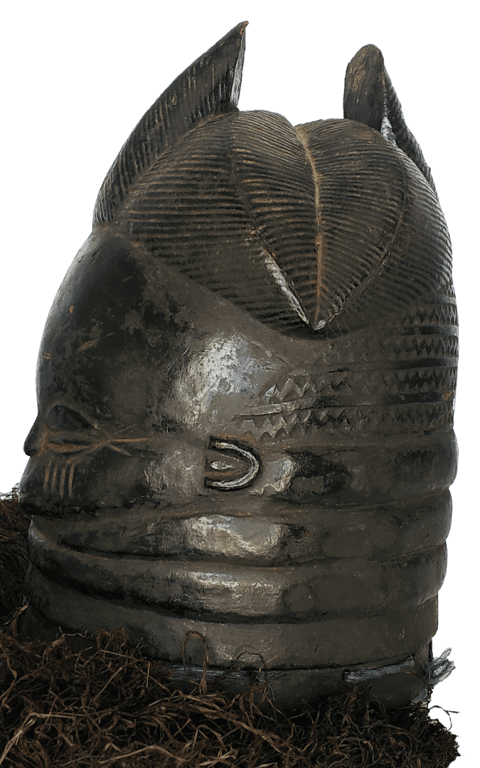 A dark, carved wooden helmet mask with geometric hair patterns and circular features, set against a soil-like texture.