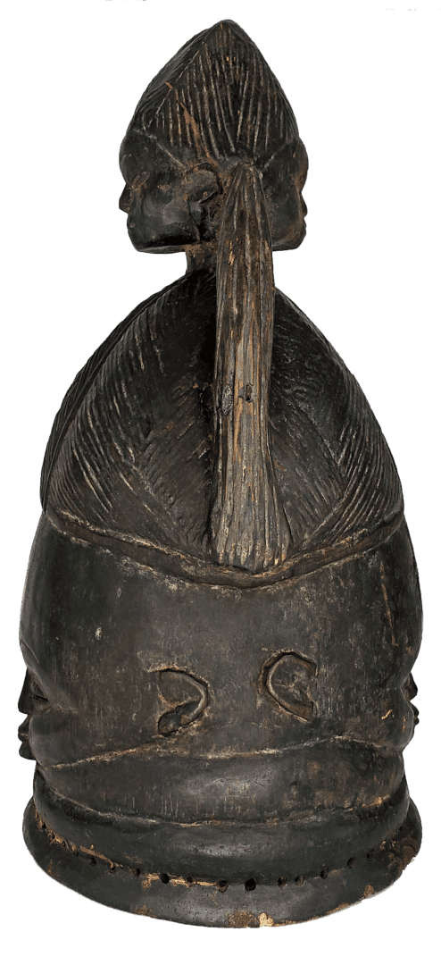 A carved, dark wooden mask depicting a human head with a central hair tuft and patterned textures, viewed from behind.