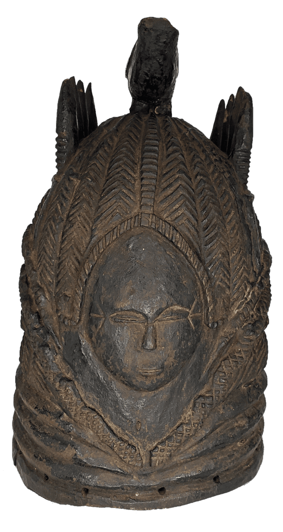 A dark, cylindrical carved wooden mask featuring a central face surrounded by intricate, feather-like textural patterns.