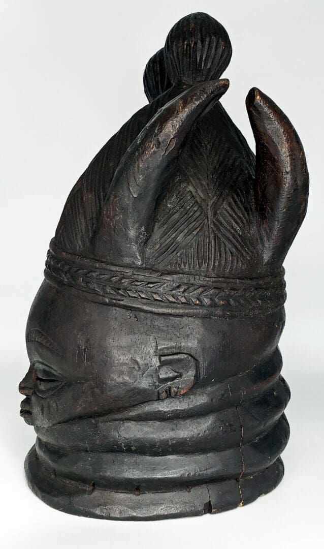 A dark wood Mende helmet mask with ringed neck, intricate braided hair, and curved, horn-like projections on the crown.