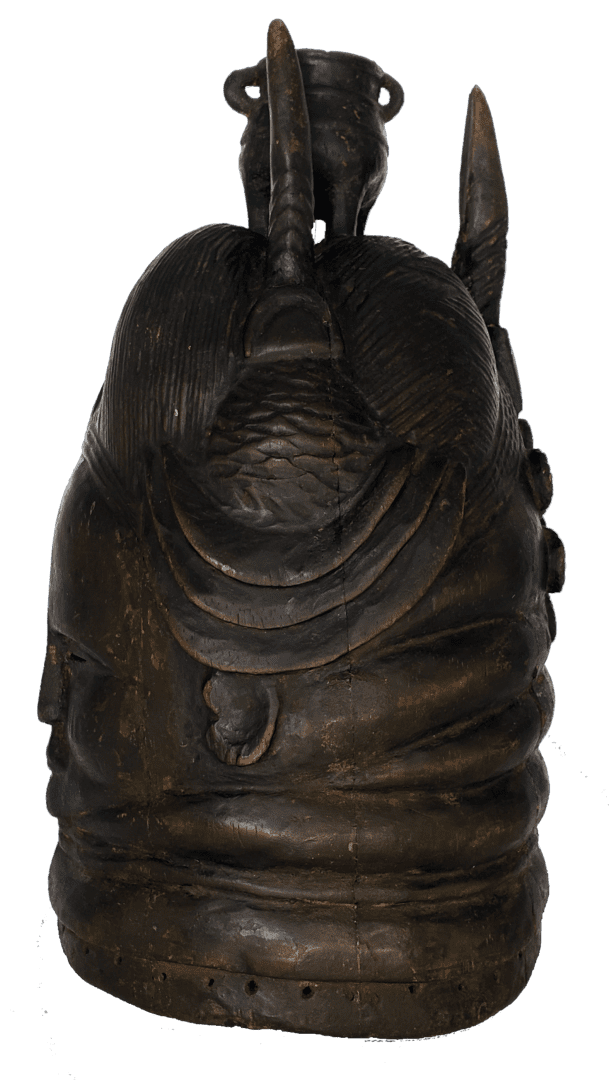A dark, carved wooden ritual helmet mask with tiered horizontal layers, topped with a small vessel and vertical motifs.