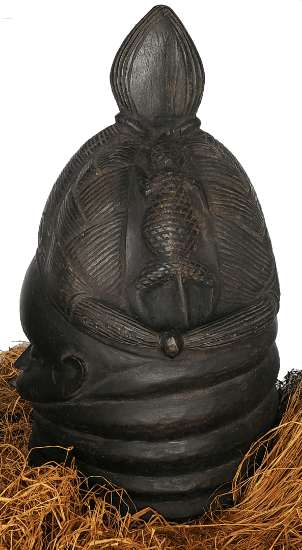A dark, carved wooden helmet mask featuring an embossed lizard motif, resting on a bed of dried straw.