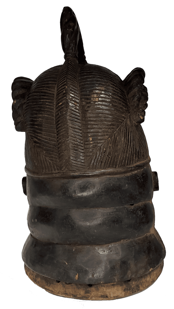 A dark, wooden, cylindrical carved mask featuring a textured, hair-like pattern on top and rounded, smooth horizontal tiers.