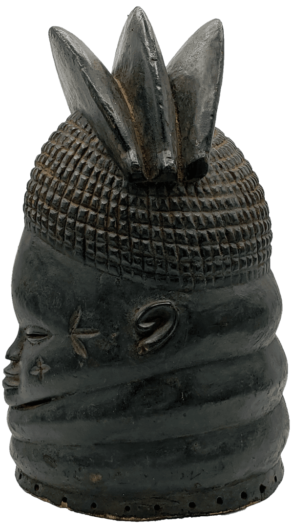 A dark, carved wooden mask with a textured cap, a triple-pointed crest, and ringed neck, set against a white background.