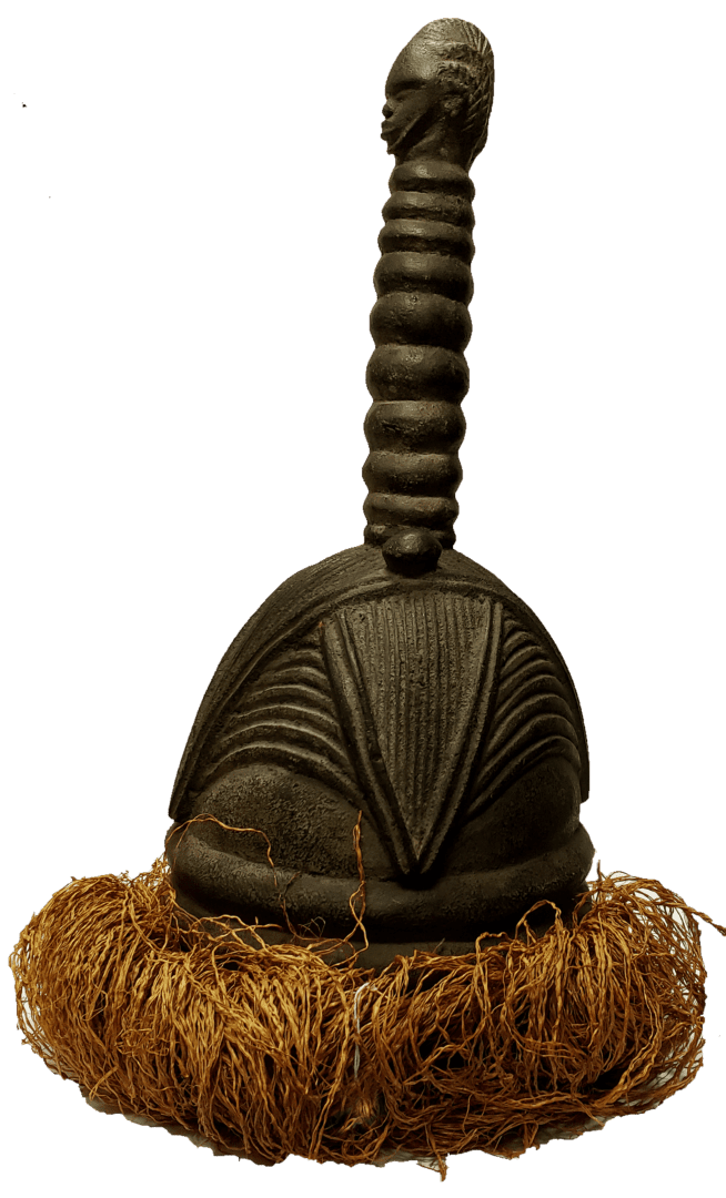 A dark, carved wooden ritual object with a long, ringed neck topped by a head, resting on a base of fibrous material.