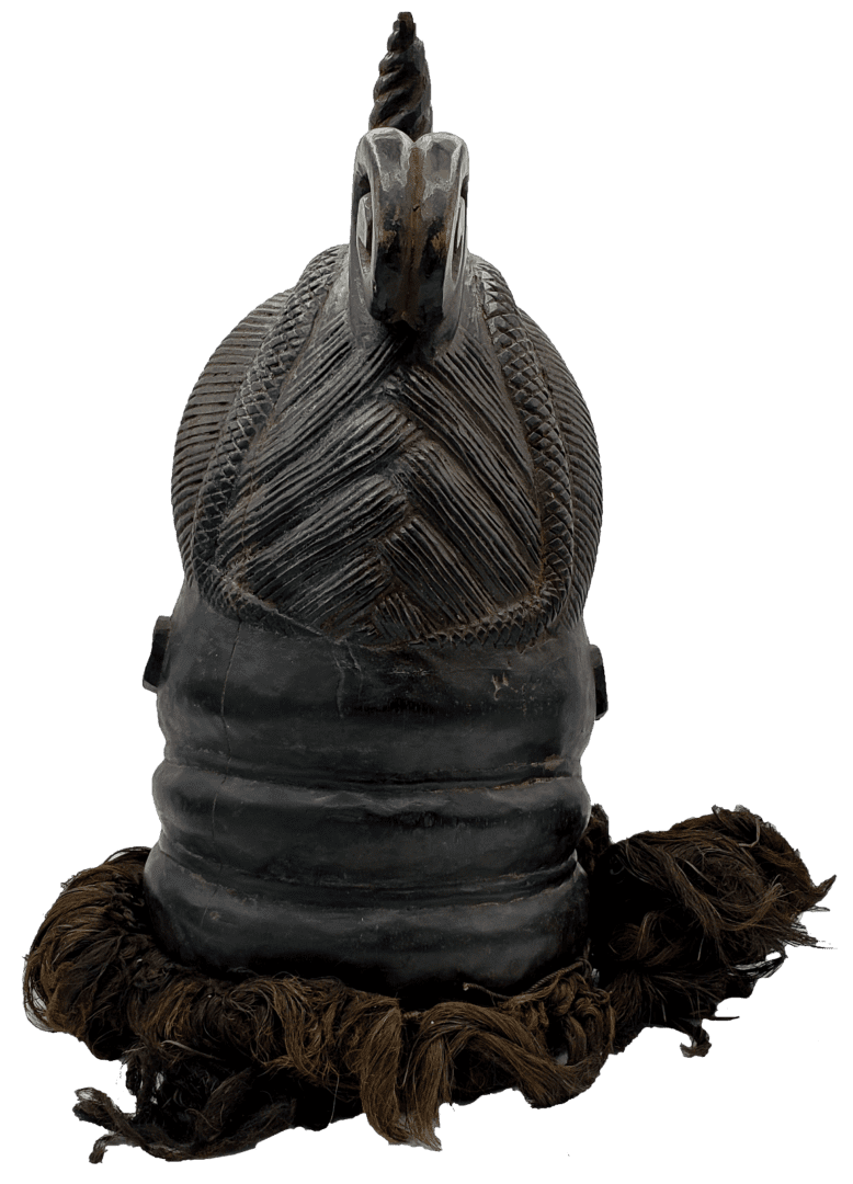 A dark carved wooden sculpture of a helmet with a textured, chevron-patterned crest and fiber fringe at the base.