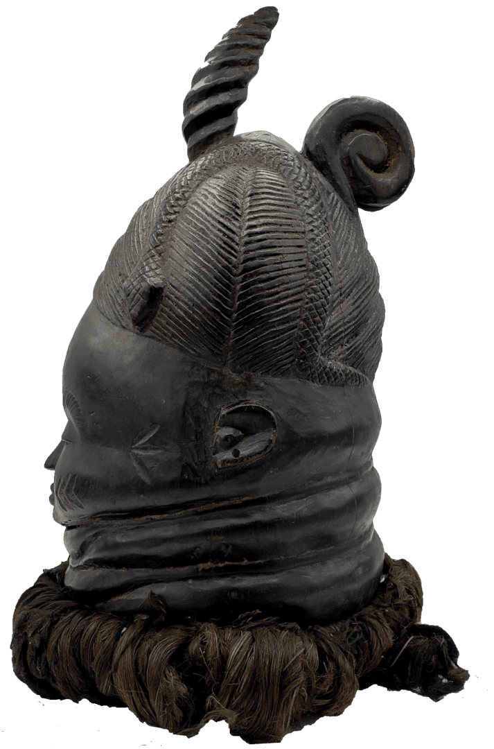 A dark, carved wooden helmet mask with intricate braided hair patterns, a spiral crest, and a thick fringe at the base.