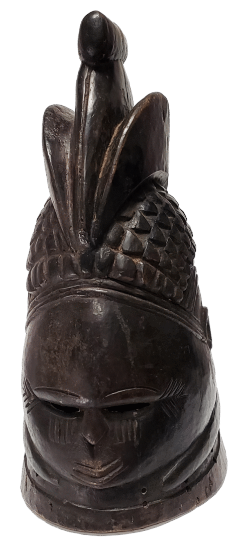 Dark carved wooden sculpture of a stylized head wearing a patterned, crown-like headdress with an upward-pointing crest.