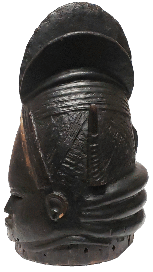 A dark, carved wooden mask with an intricate, crested hairstyle and horizontal neck rings, seen from a side-rear angle.
