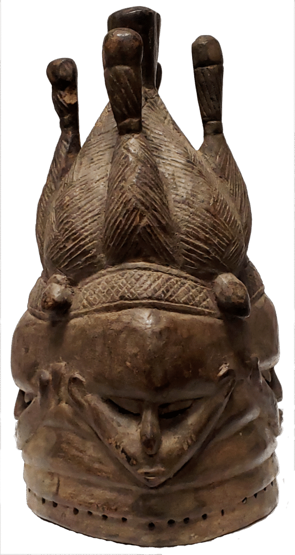 A brown, cylindrical carved wood mask featuring a human face, elaborate textured hairstyle, and several small figures.