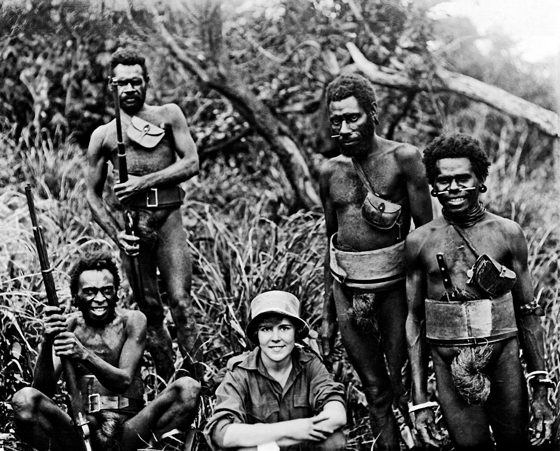A woman in a sun hat sits amongst four men carrying rifles in a dense, brush-filled environment.