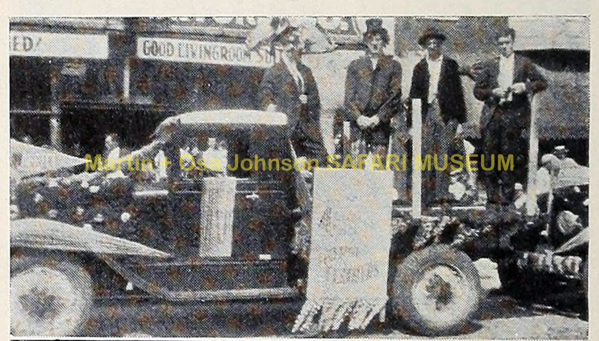 A vintage truck carrying four people stands in a street, with a banner draped over the side and a museum watermark.