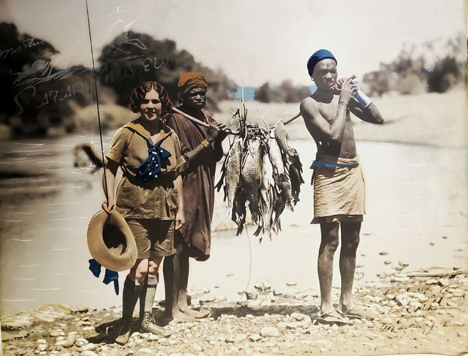 A woman and two men stand on a riverbank, holding a fishing rod and a large string of fish.