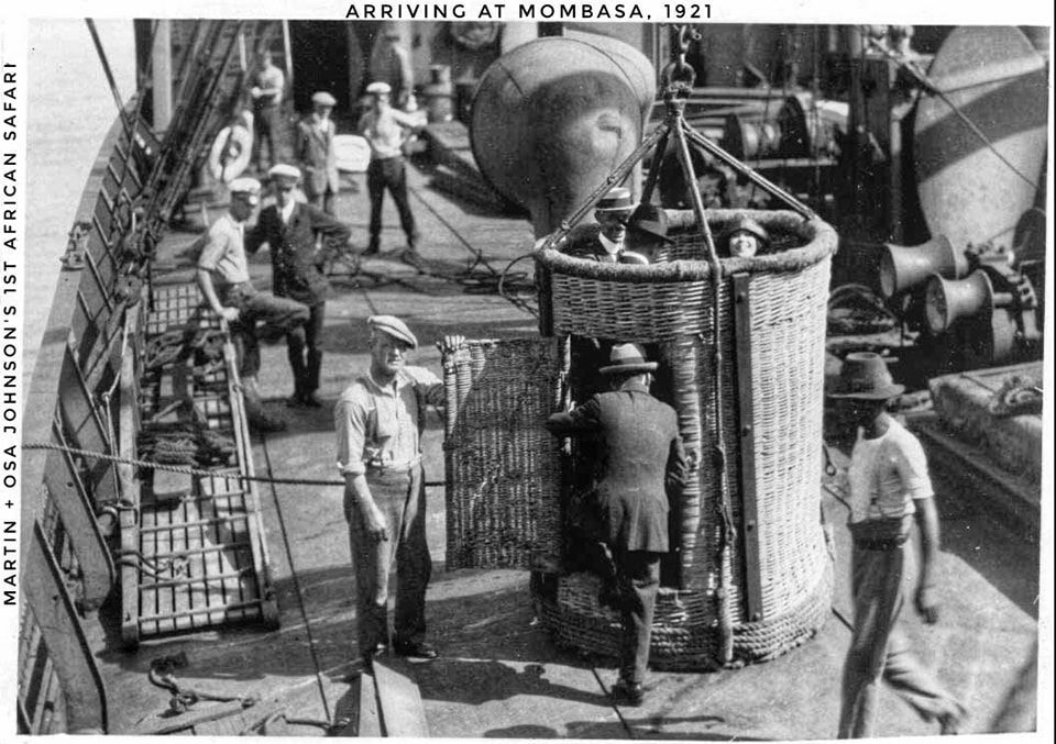 People in hats and suits use a wicker crane basket on a ship deck labeled
