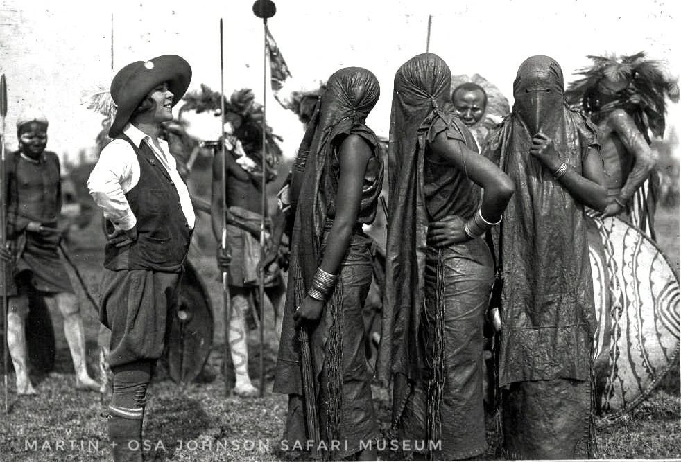 A person in Western safari attire stands before a group of people wearing traditional ceremonial veils and holding shields.