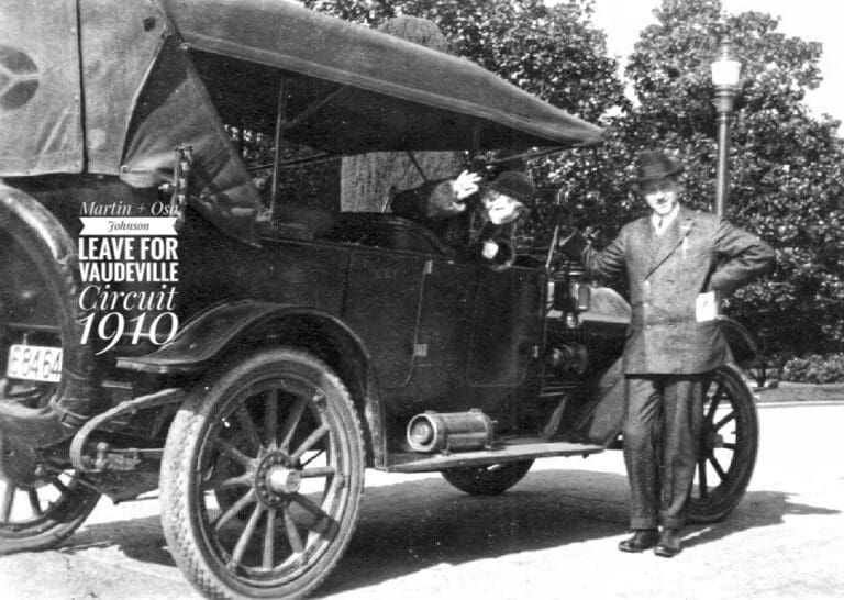 A black-and-white photo shows a man standing beside a vintage car with someone waving from the driver's seat.
