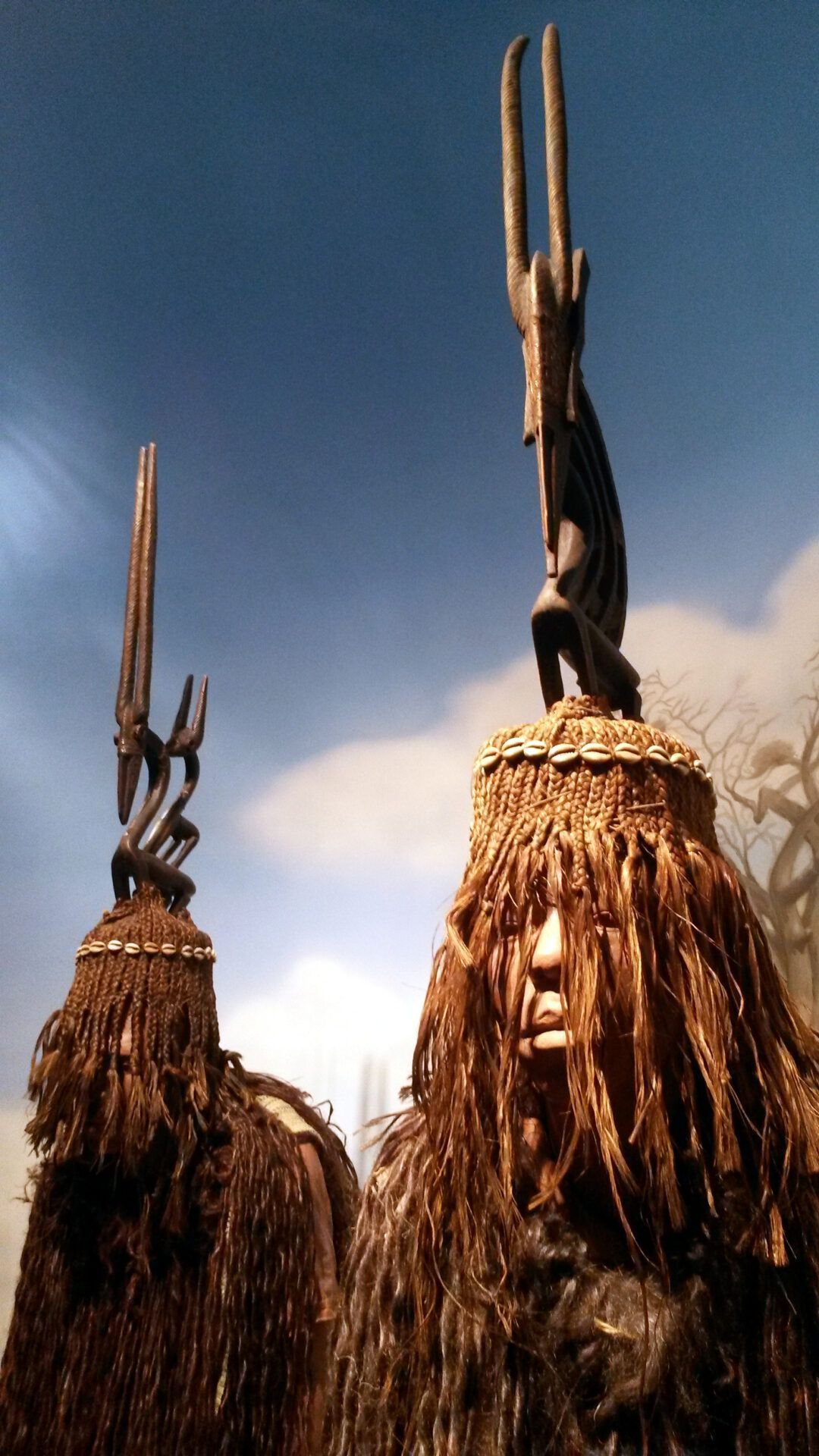 Two figures in dark, fibrous costumes with tall, sculpted wooden headdresses stand against a light blue sky.