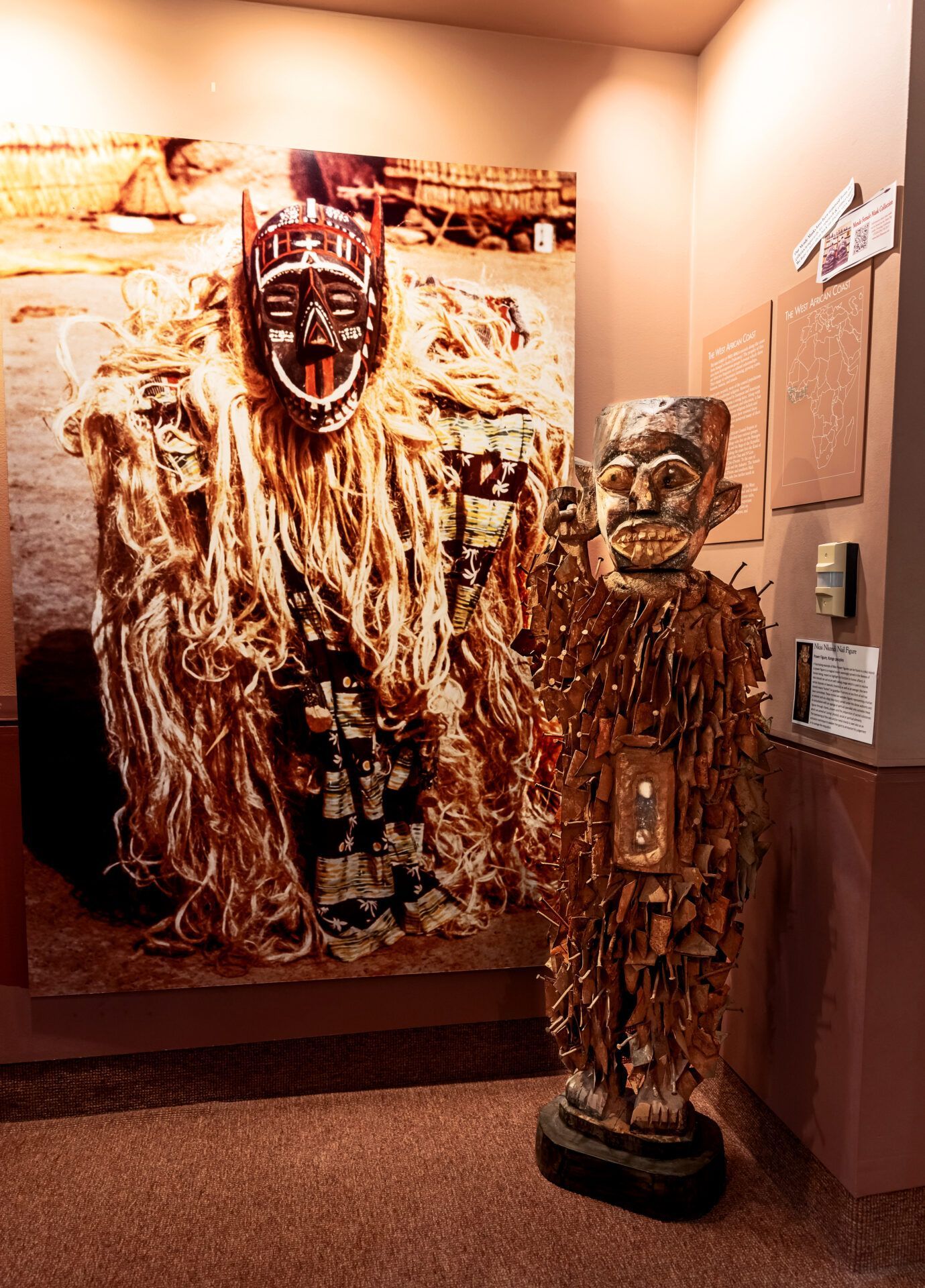 A museum display featuring a large wall photograph of a masked figure alongside a three-dimensional figure in costume.