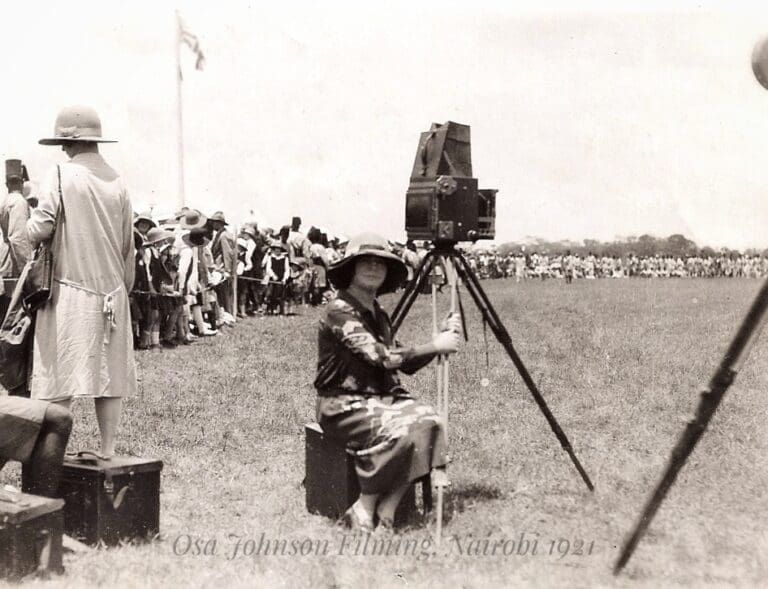 Osa Johnson seated on a suitcase while filming with a large vintage movie camera on a tripod in Nairobi, 1921.