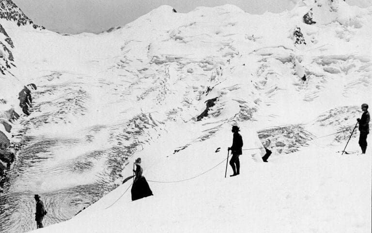 A historical black-and-white photograph of a group roped together trekking across a snowy, mountainous glacial landscape.