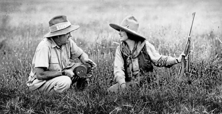 Two people in outdoor attire and brimmed hats crouch in a field, communicating while one holds a rifle.