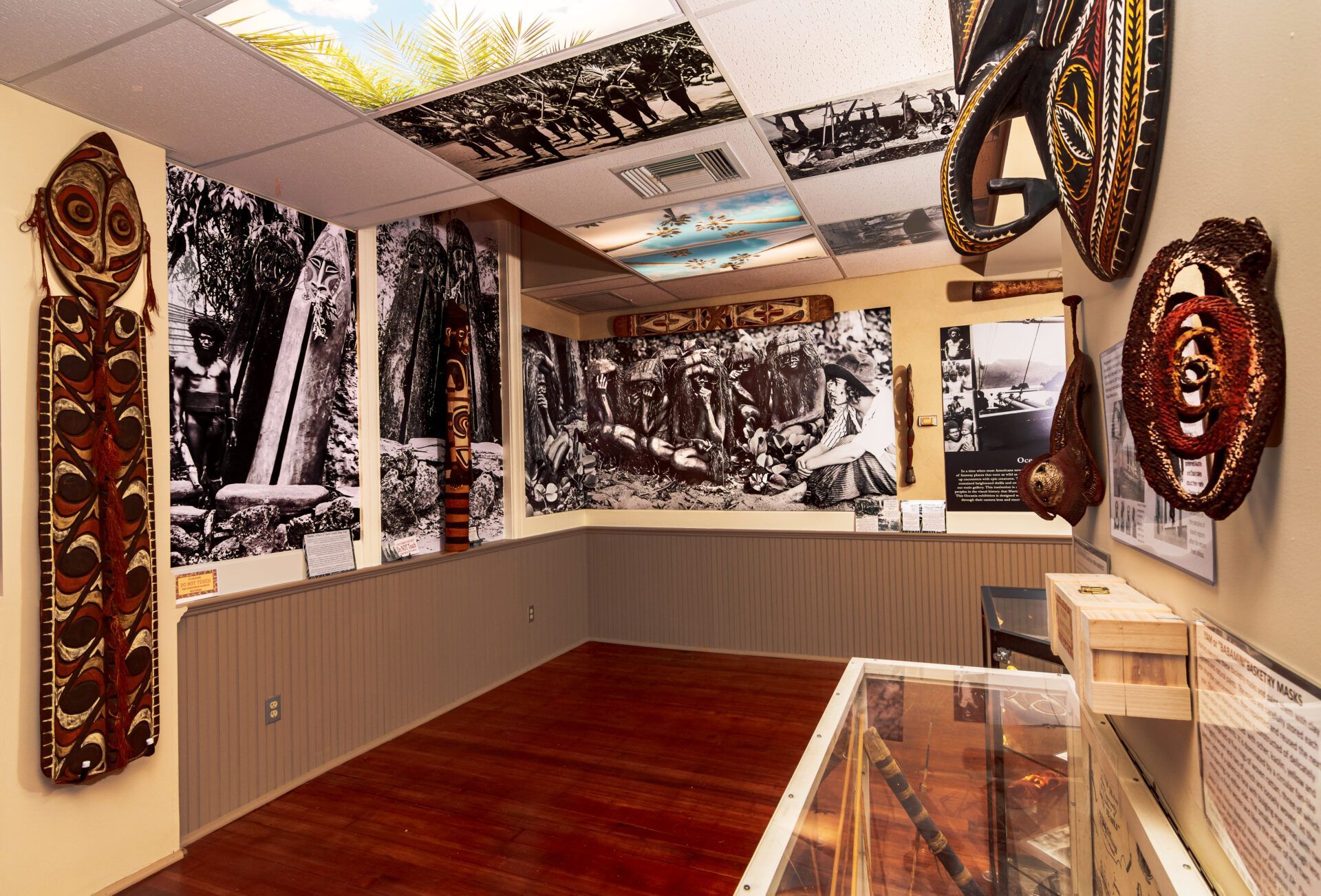 An indoor museum exhibit displays tribal wooden masks and black-and-white historical photographs on the walls and ceiling.
