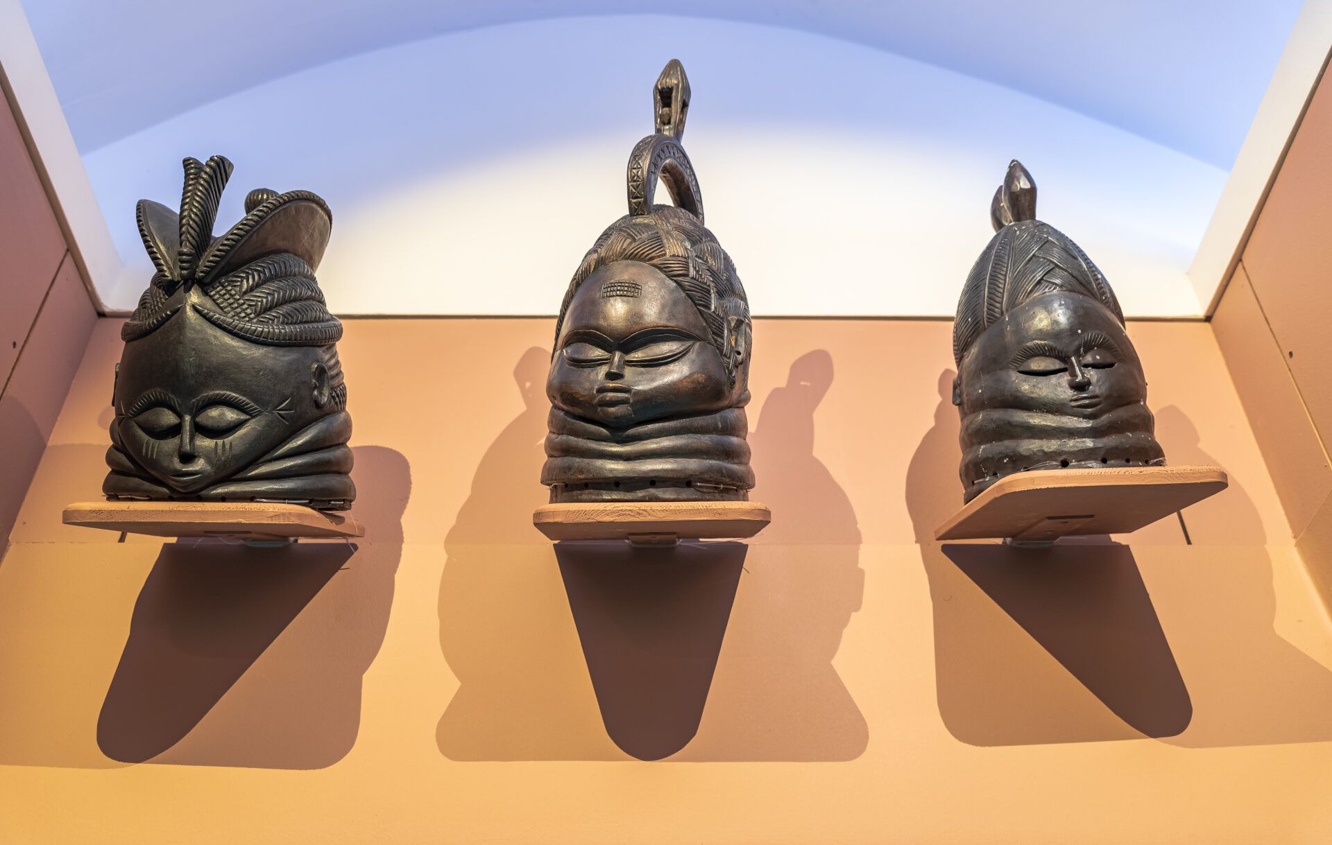 Three dark-toned, carved sculptural masks displayed on wall-mounted shelves against a light tan wall.