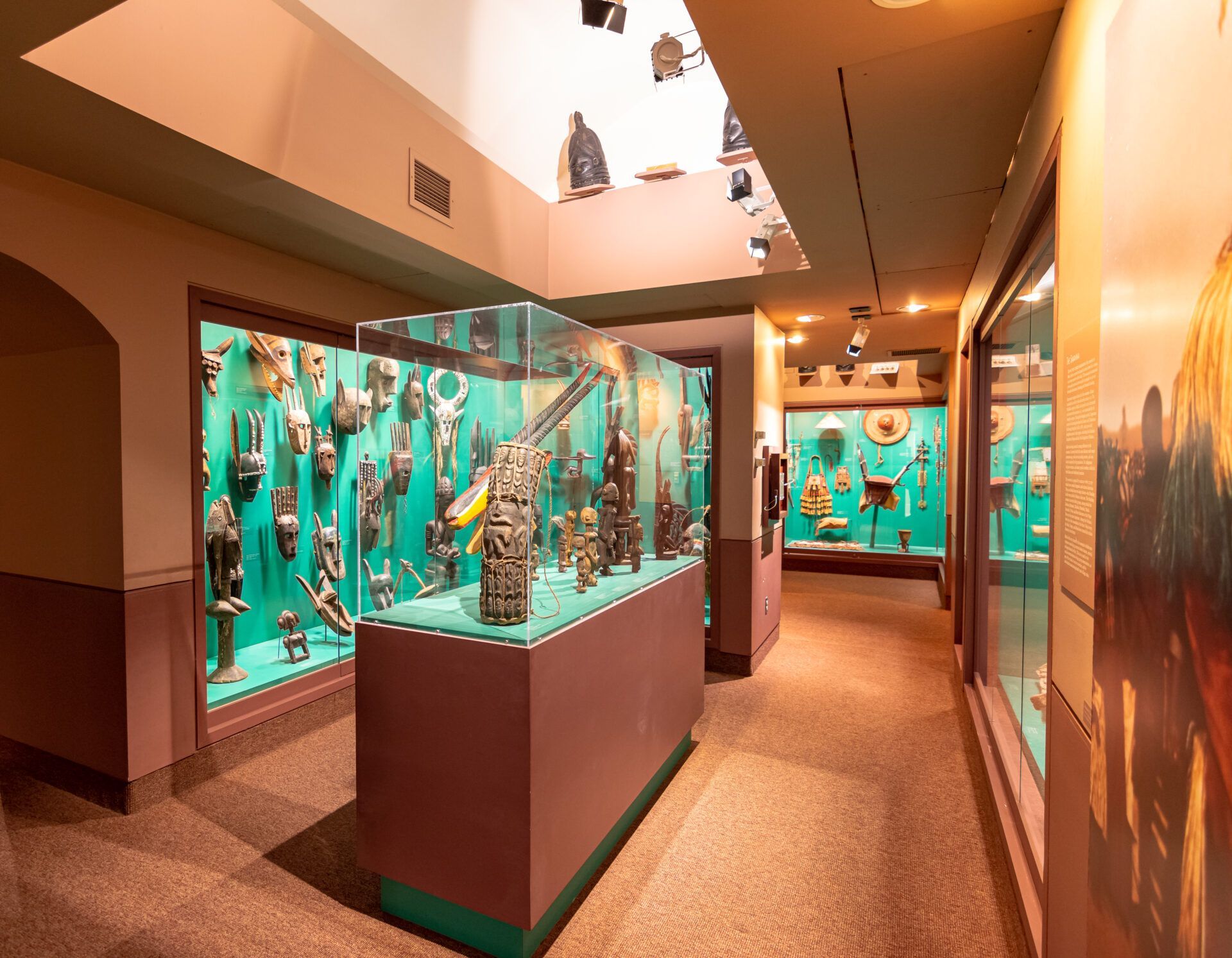 A museum exhibit hallway with teal-backed glass display cases containing numerous carved masks and artifacts.