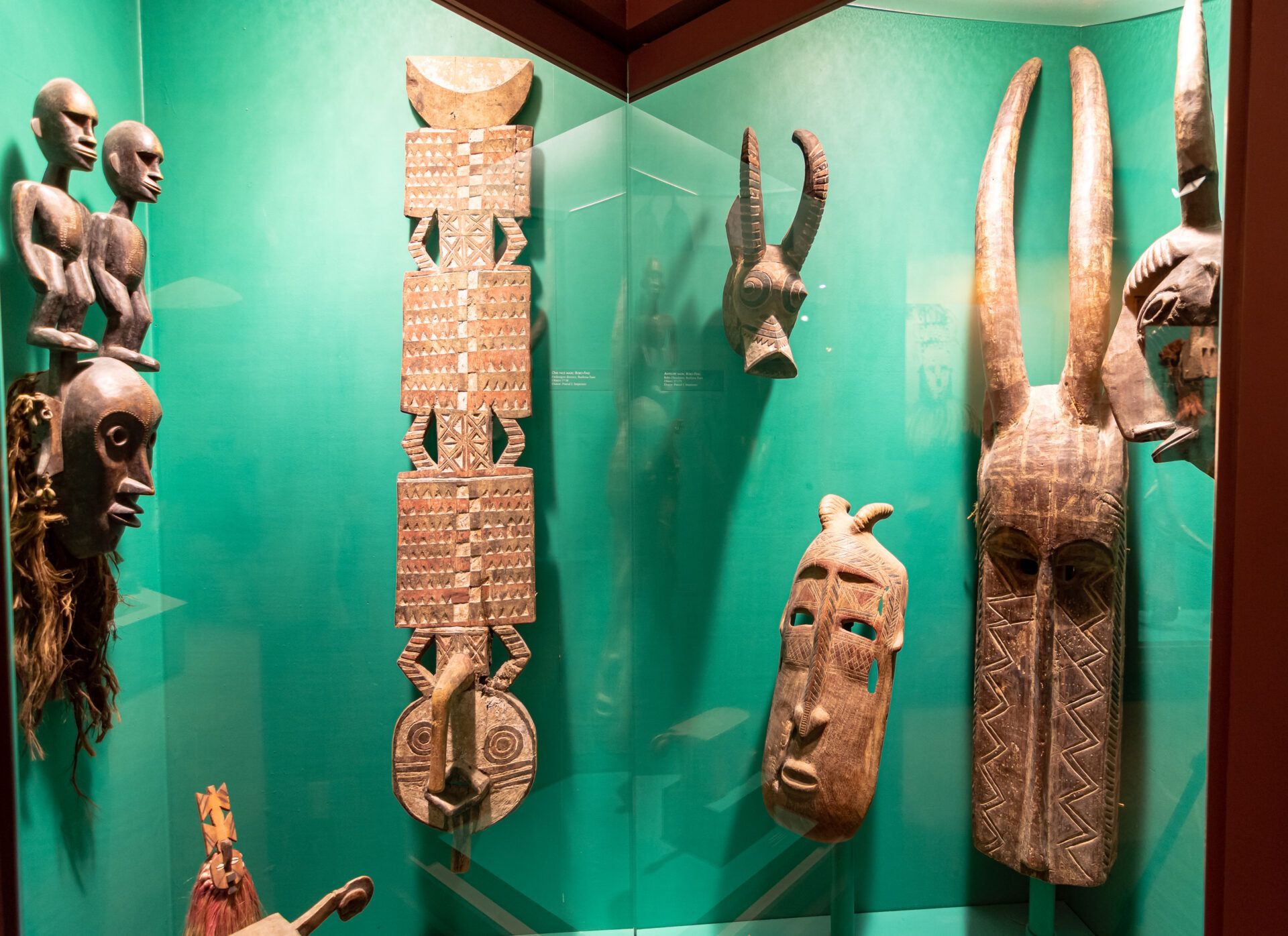 Display of traditional wooden sculptures and masks with carved patterns, set against a teal background in a museum case.