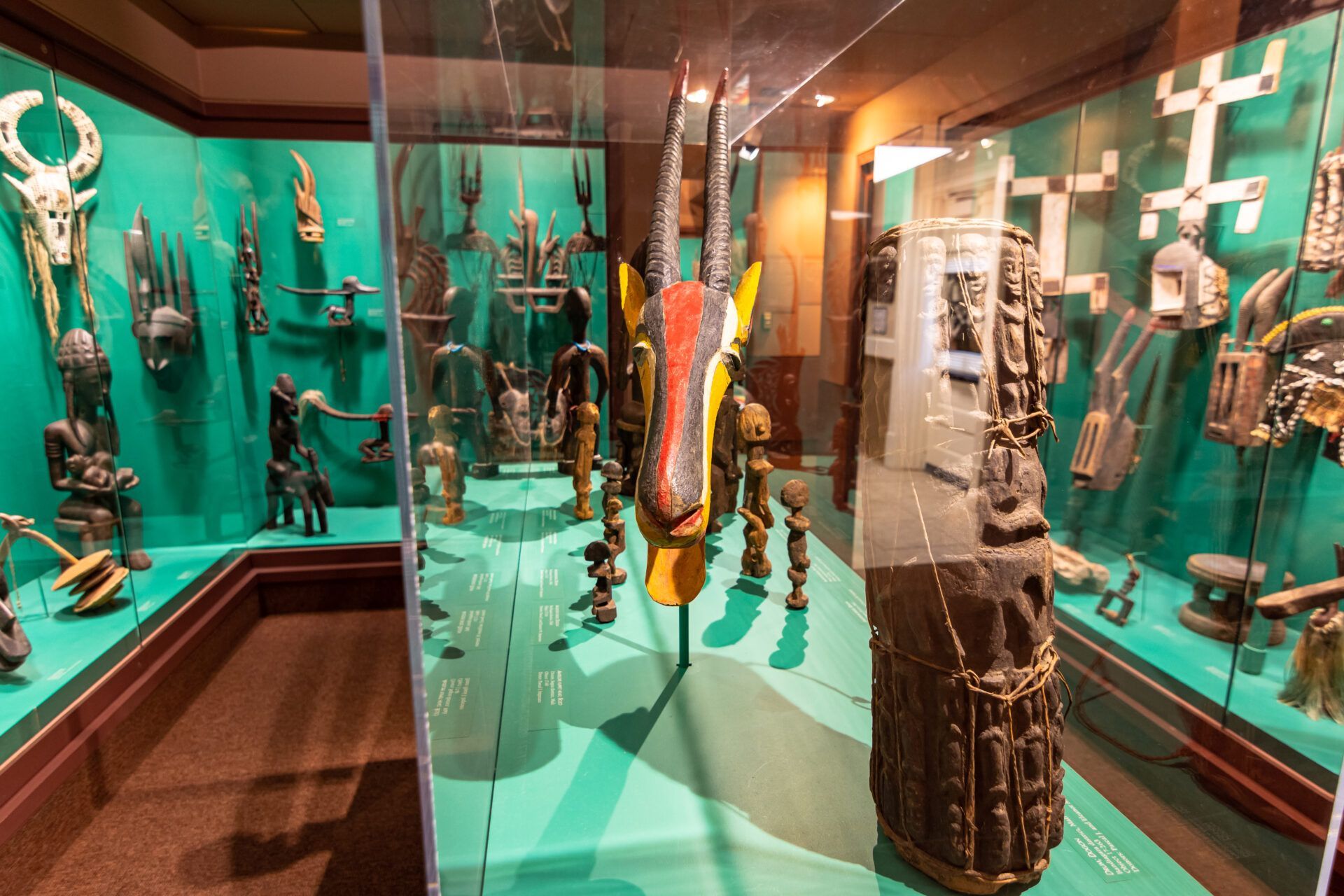 Museum display cases filled with diverse tribal masks and wooden sculptures against a vibrant teal background.