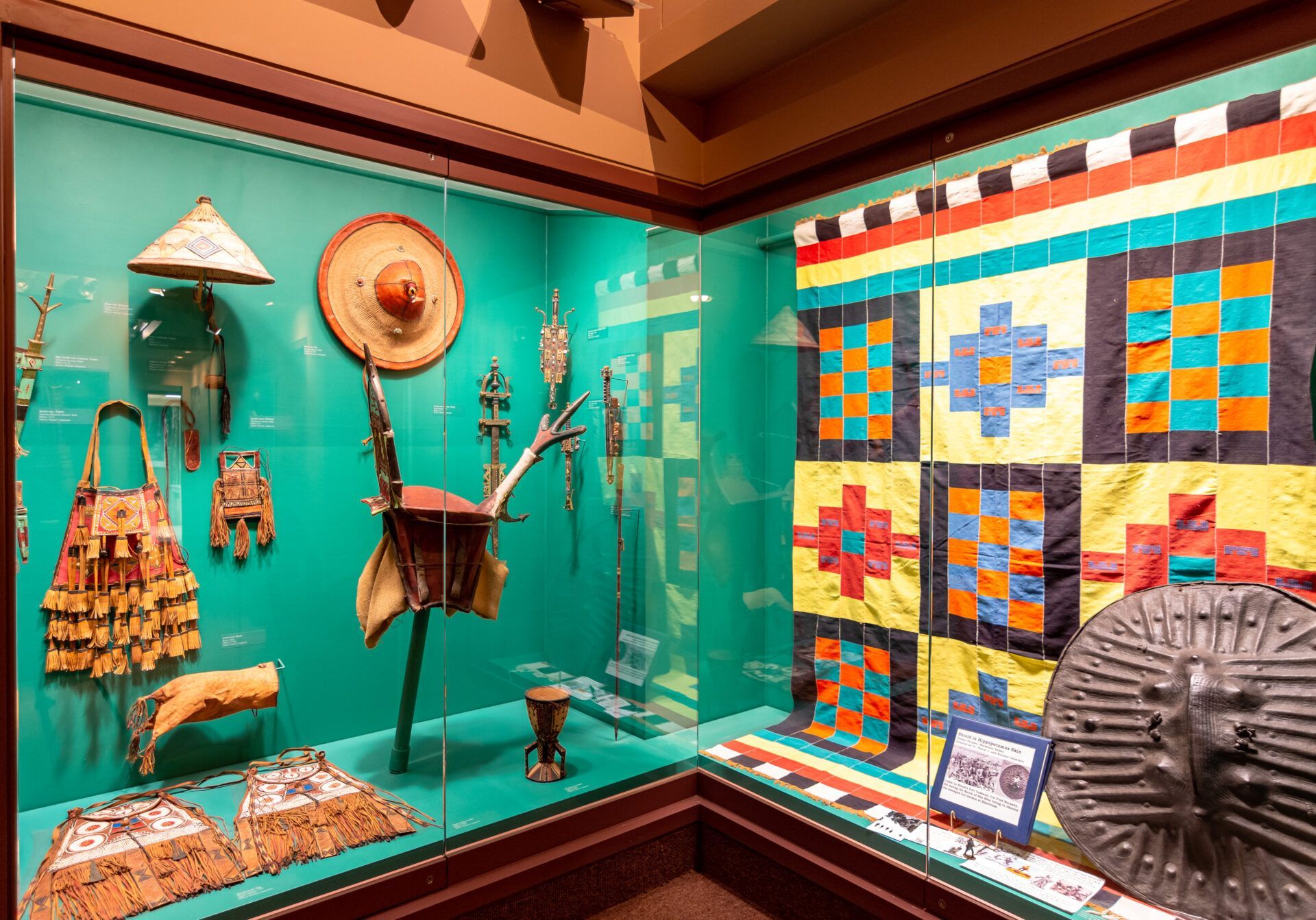A museum display case containing traditional indigenous artifacts, including garments, a geometric quilt, and a shield.
