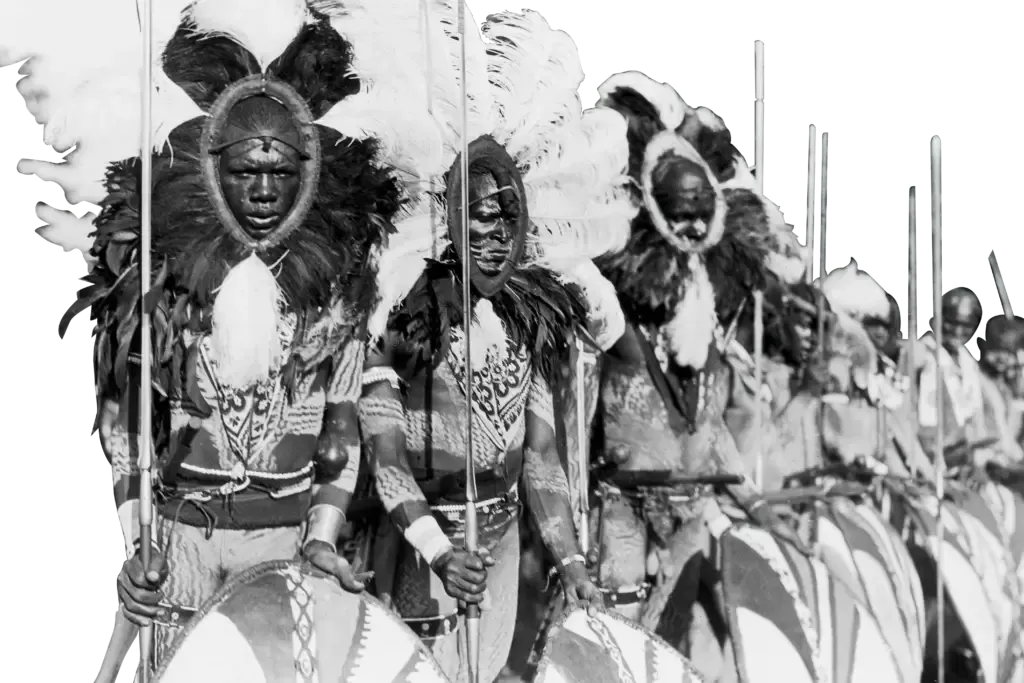 A row of people wearing traditional headdresses and ornate patterned clothing, holding spears and large, circular shields.