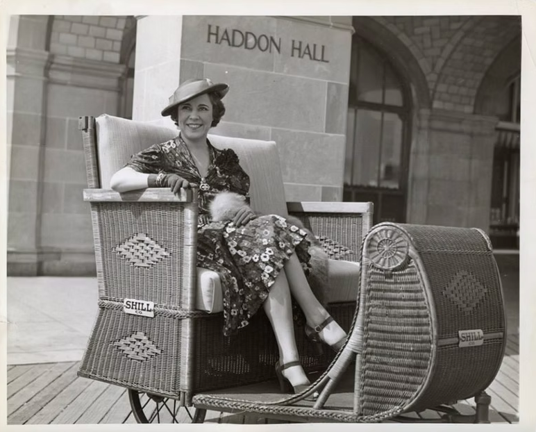 A smiling person sits in a wicker rolling chair on a wooden boardwalk in front of a building labeled Haddon Hall.