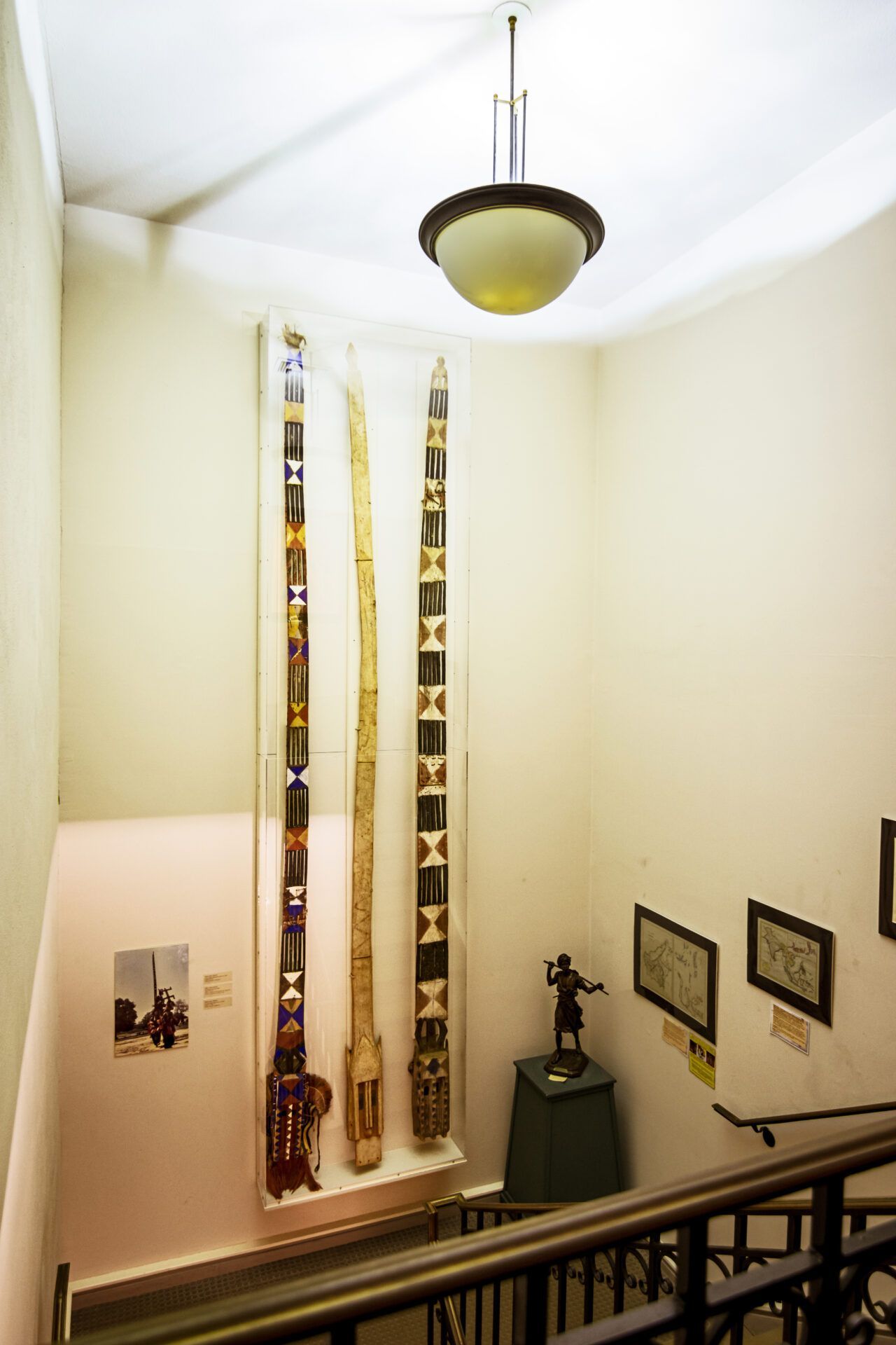 A museum staircase with three long decorative poles mounted on a wall, a light fixture above, and small wall art below.