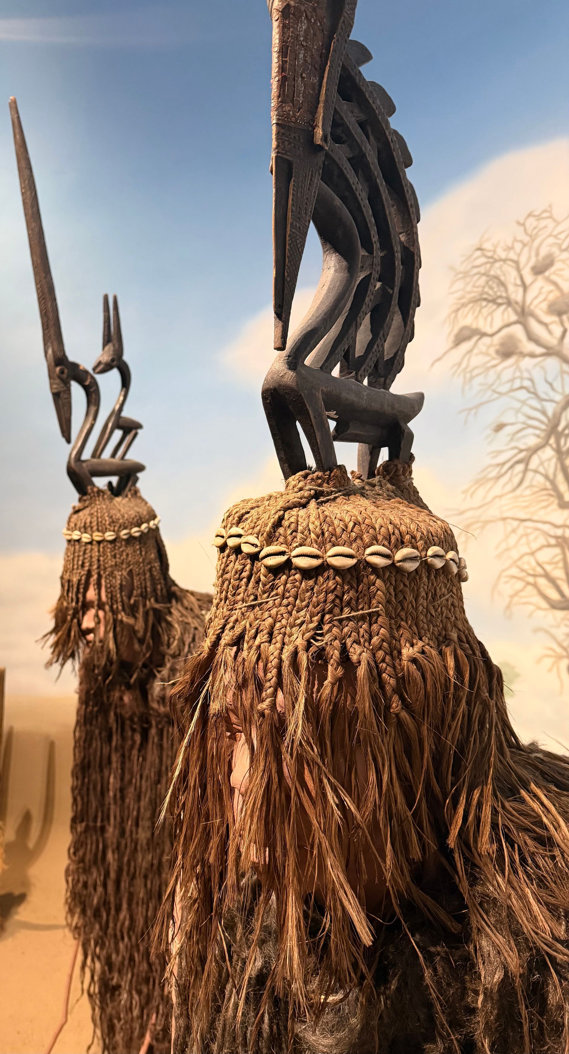Two carved wooden figures atop straw masks, decorated with cowrie shells and long, dark, fibrous fringes.